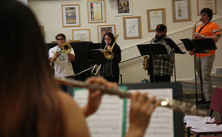 A closeup of a flautist playing their instrument across the room from a group of four horn players who are also playing theirs. They are mixed in terms of gender and race and dressed in casual clothing.
