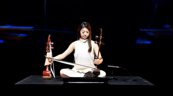 Instrumentalist Jeonghyeon Joo playing a haegeum while cross-legged on a platform. She and Asian woman with long dark hair, dressed in white clothing with her arms uncovered.